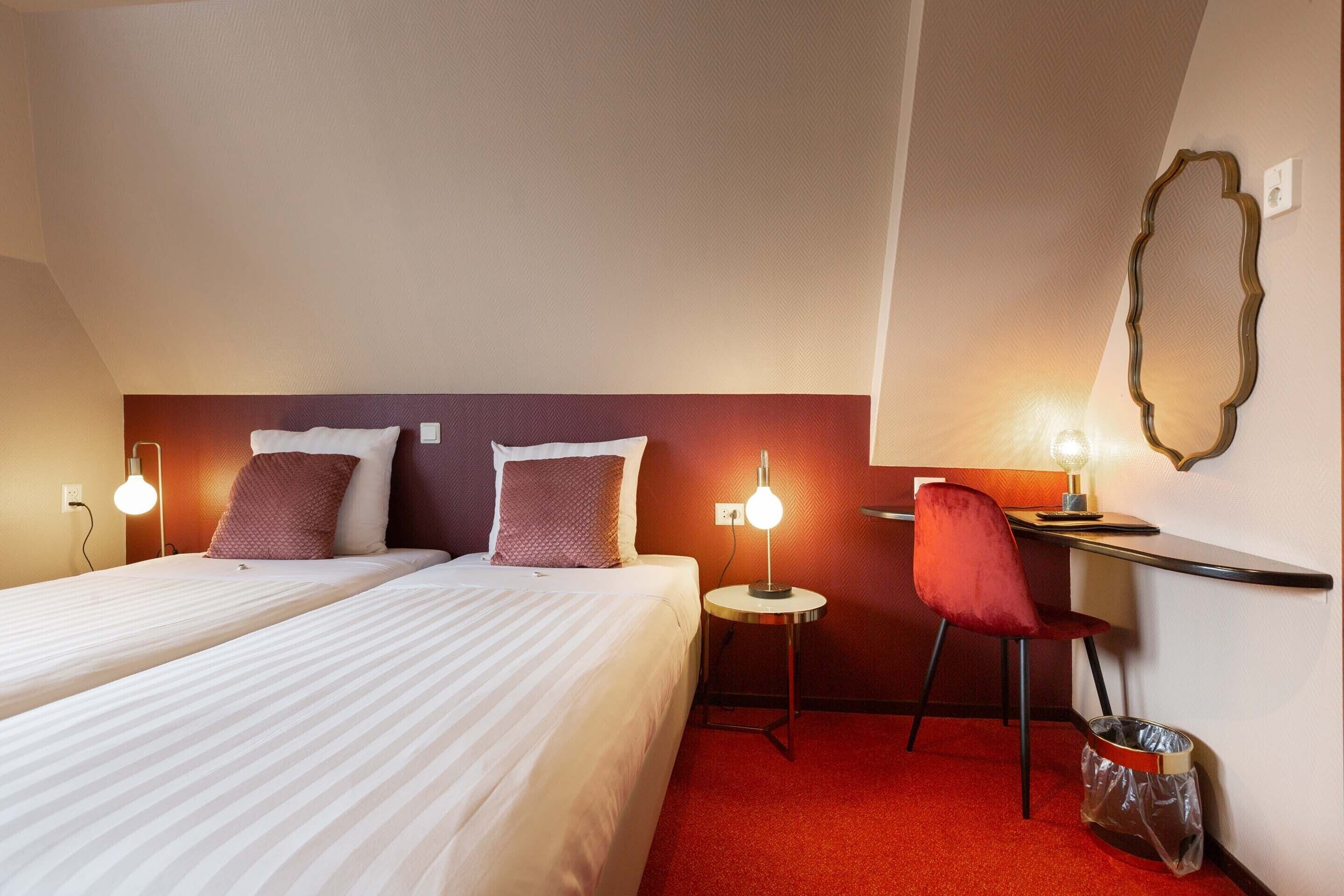 A hotel room with two beds, a desk, a chair, and a trash bin.