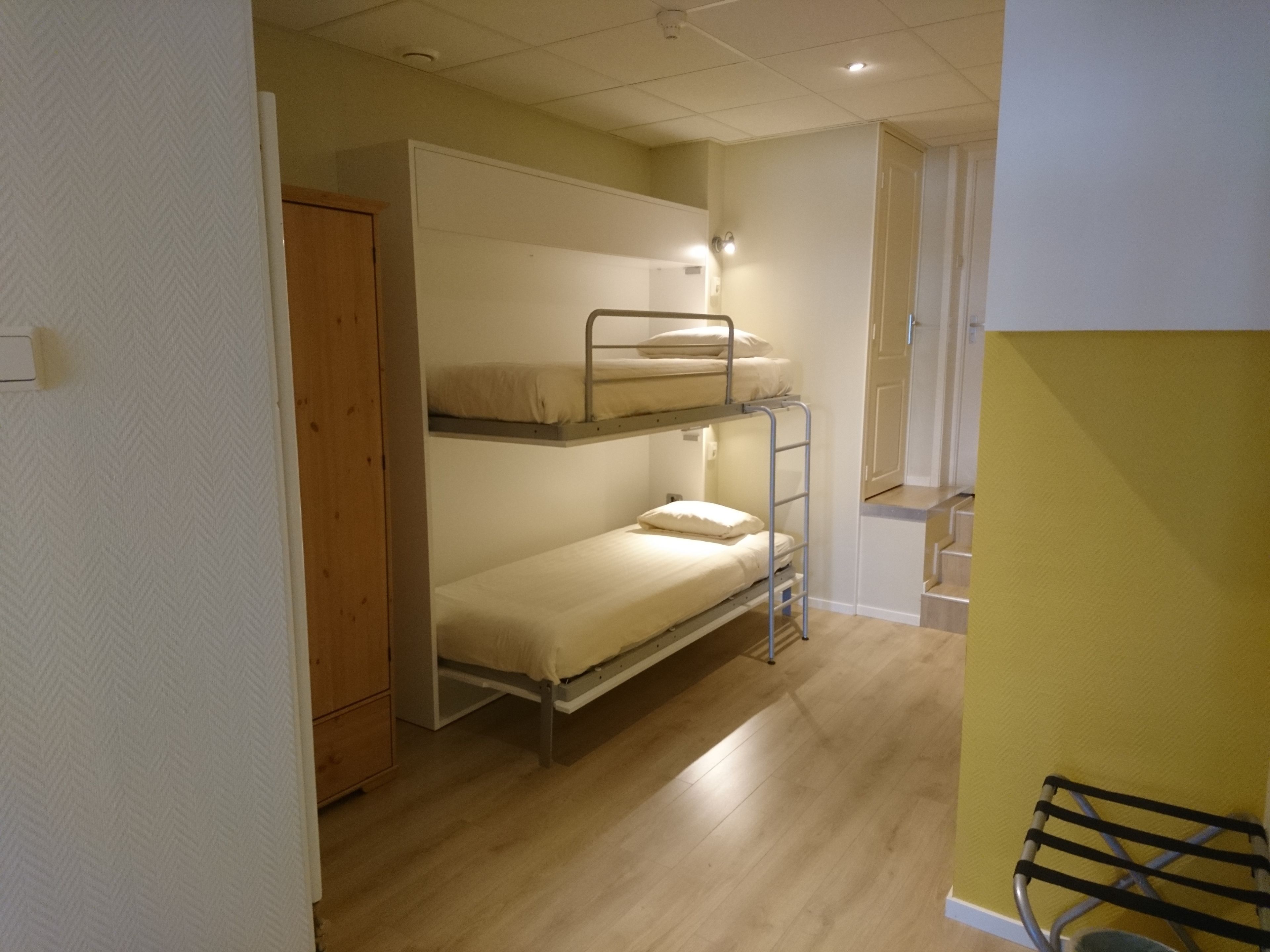 Quarto em dormitório com beliches, piso de madeira e uma parede amarela de destaque.