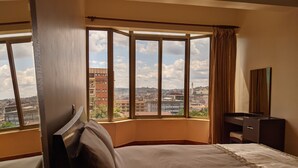 Une chambre d’hôtel dotée d’une grande fenêtre donnant sur la ville, d’un lit, d’une commode et d’un miroir.