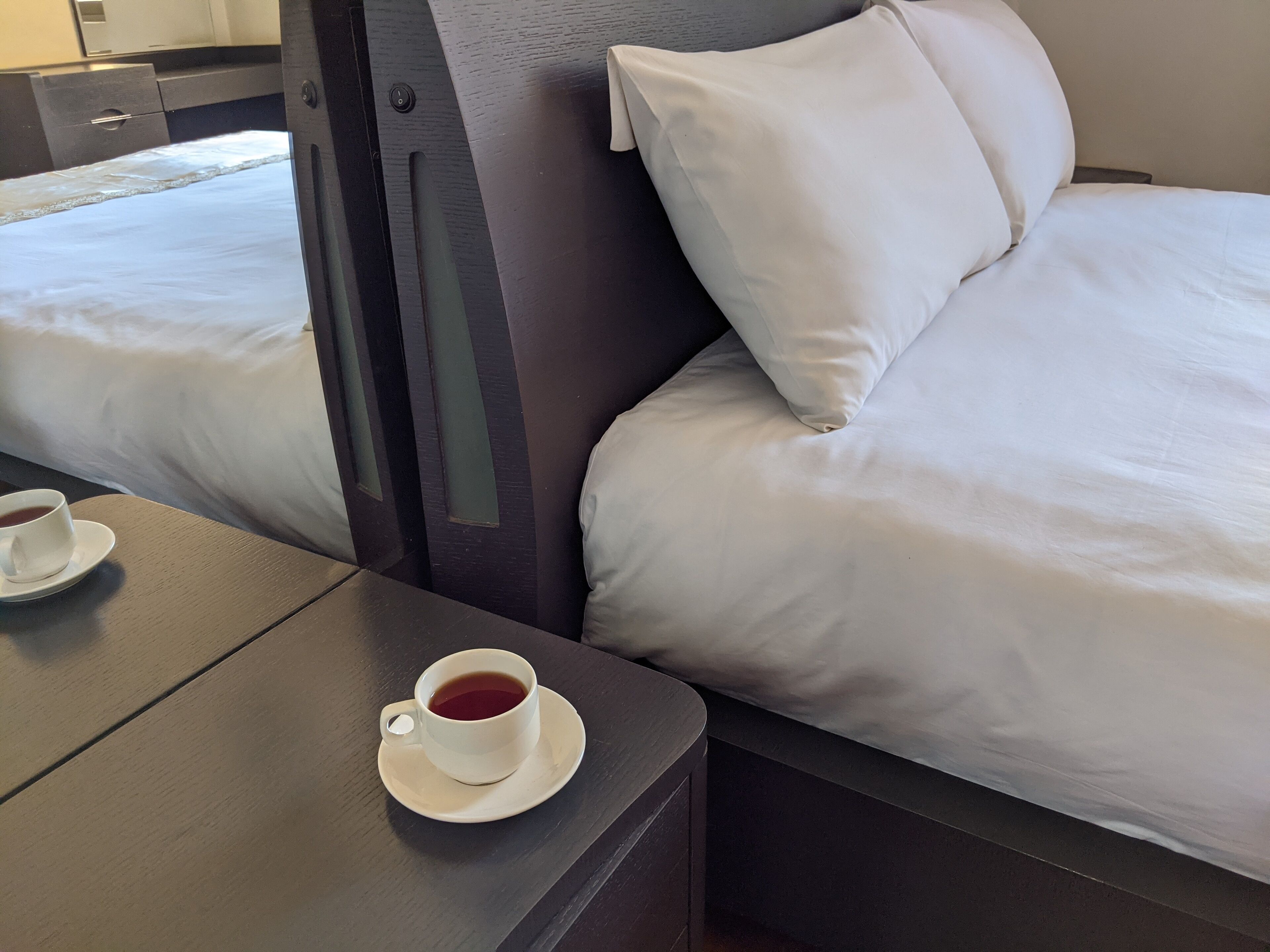 A hotel room with two beds, a wooden nightstand with a cup of tea, and a dark-colored wardrobe.