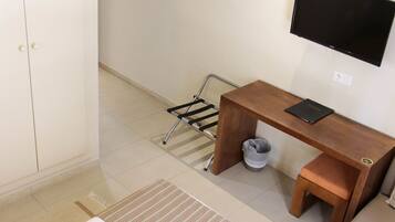 A small, clean room with a wooden desk, a TV mounted on the wall, a trash bin, and a metal rack.