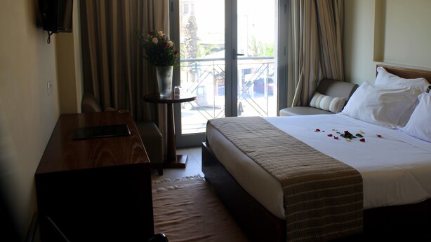 A hotel room with a bed, a desk, a chair, a TV mounted on the wall, and a balcony with curtains. - Résidence Le Consul (Tunis)