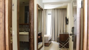 A narrow hallway leading to a bedroom with a bed, a small stool, and a chair. A mirror and a television are mounted on the wall. - Résidence Le Consul (Tunis)