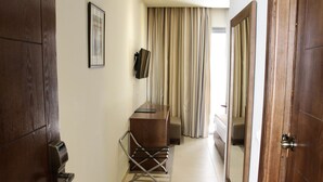 A hotel corridor with a wooden door, a desk with a chair, a television mounted on the wall, and a large window with curtains. - Résidence Le Consul (Tunis)
