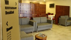 A hotel lobby with grey sofas, a wooden partition, a TV, and a sign indicating the restaurant and breakfast area. - Résidence Le Consul (Tunis)