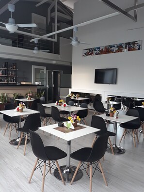 A modern dining area with multiple tables and chairs, a kitchen in the background, and a flat-screen TV mounted on the wall. - Victoria Hotel (Castello di Cisterna)