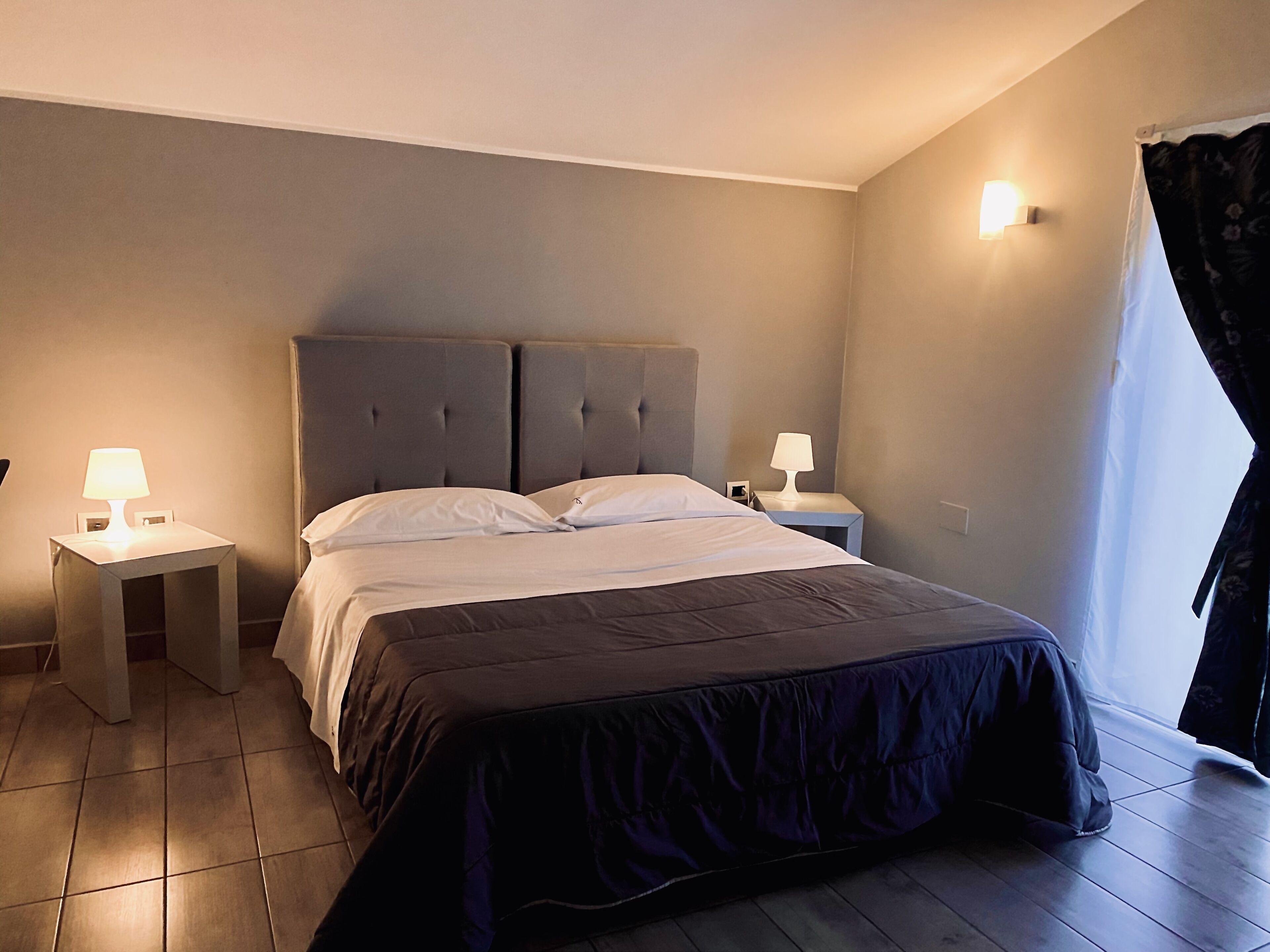 A bedroom with a bed, two bedside tables, a nightstand, a wall-mounted lamp, and a window with curtains.