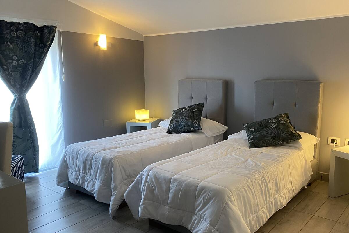 A hotel room with two beds, a nightstand with a lamp, a window with curtains, and a wall-mounted light fixture.