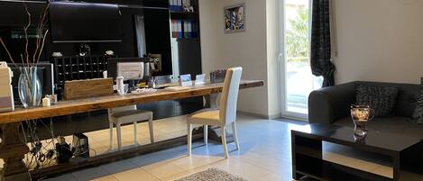 Uma sala de estar moderna com uma mesa de jantar de madeira, cadeiras brancas, um sofá preto e uma mesa de centro. Há uma TV fixada na parede, uma estante e uma janela com cortinas.