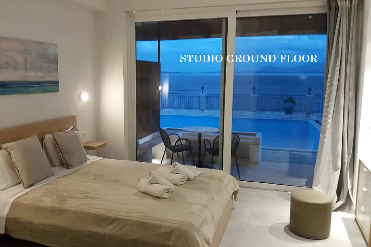 A hotel room with a bed, a nightstand, a bedside table, a chair, a small round ottoman, and a balcony with a pool and chairs.