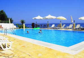 A pool area with lounge chairs and umbrellas, a clear blue sky, and a view of the sea. - Oceanis Rooms Apartments (Corfu)