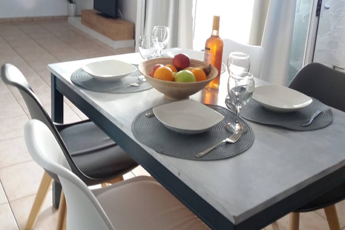 A dining area with a table set for two, a bowl of fruit, and a bottle of wine.