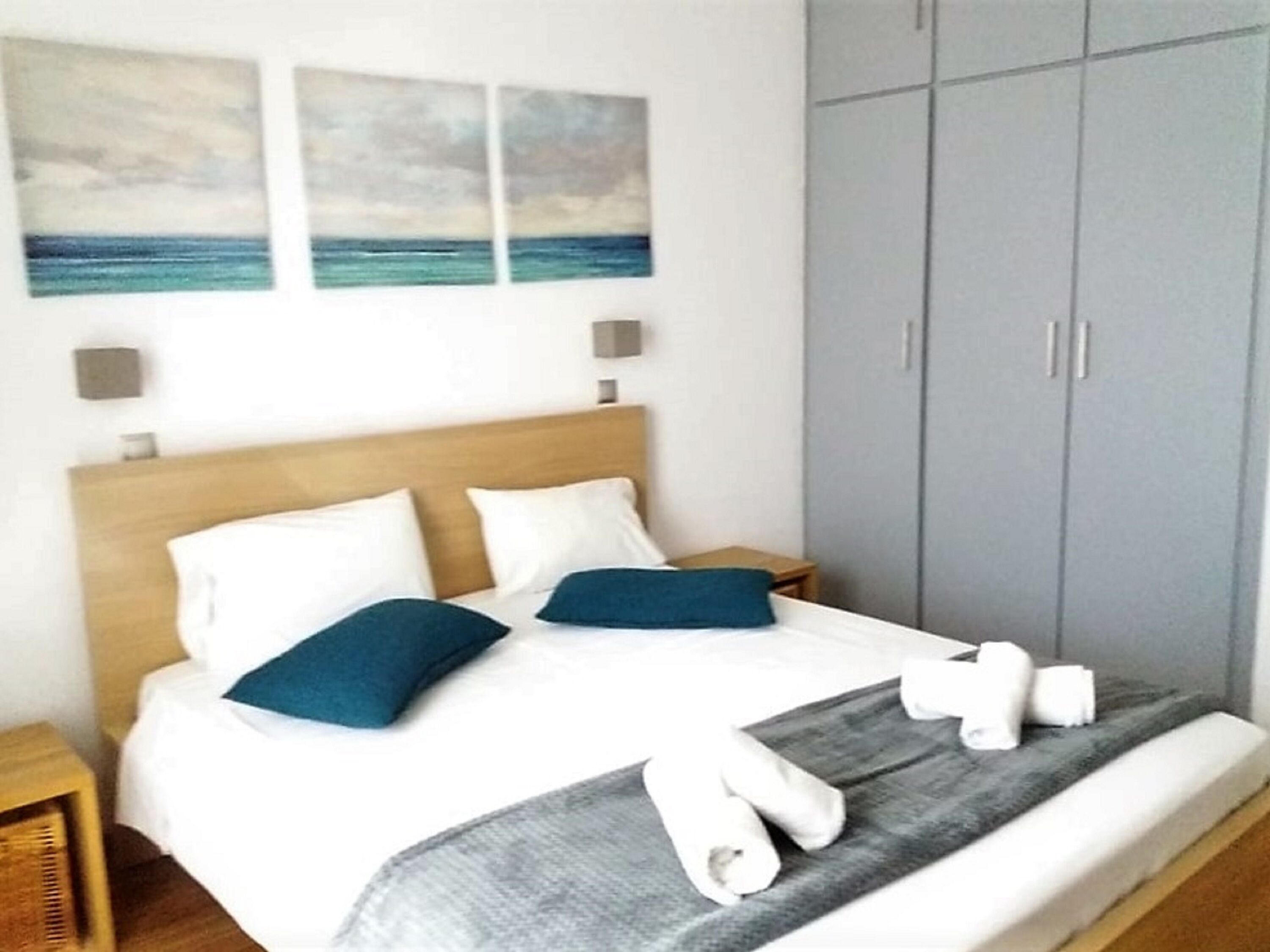 A neatly made bed with white and blue pillows, a grey blanket, and rolled towels on a wooden headboard. There are two wall-mounted light fixtures and a grey wardrobe to the right.