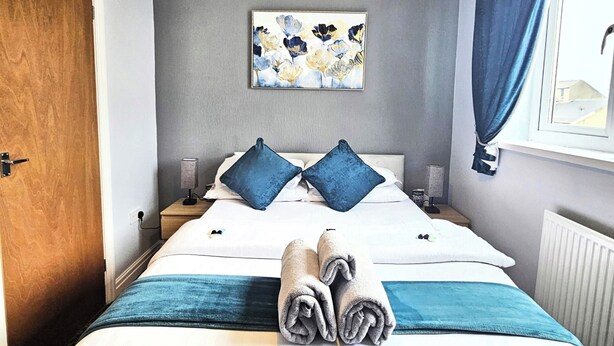 A hotel room with a bed, two pillows, a blue blanket, a rolled towel, a painting on the wall, a window with curtains, and a lamp on the nightstand. - Denville Hotel (Blackpool)