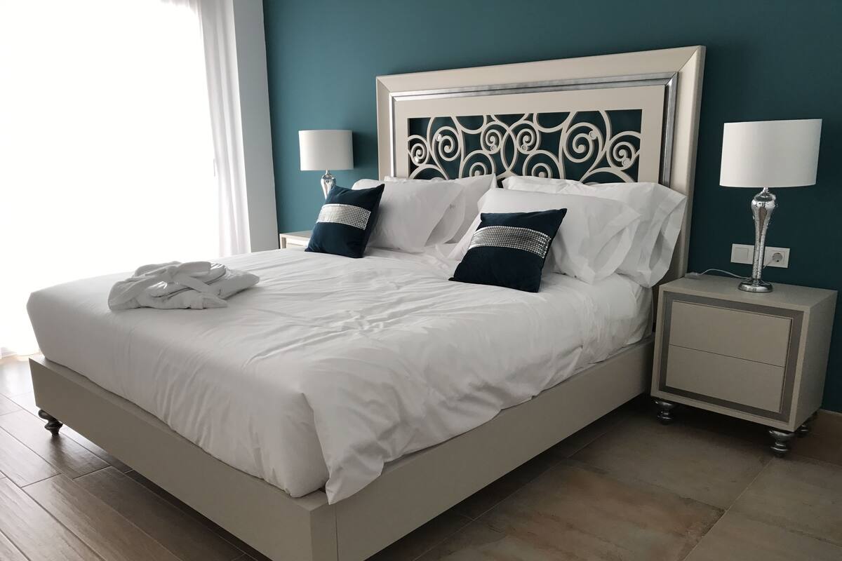 Un lit bien fait, avec du linge de lit blanc et des coussins décoratifs, une table de chevet avec une lampe et une petite table de nuit également dotée d’une lampe.