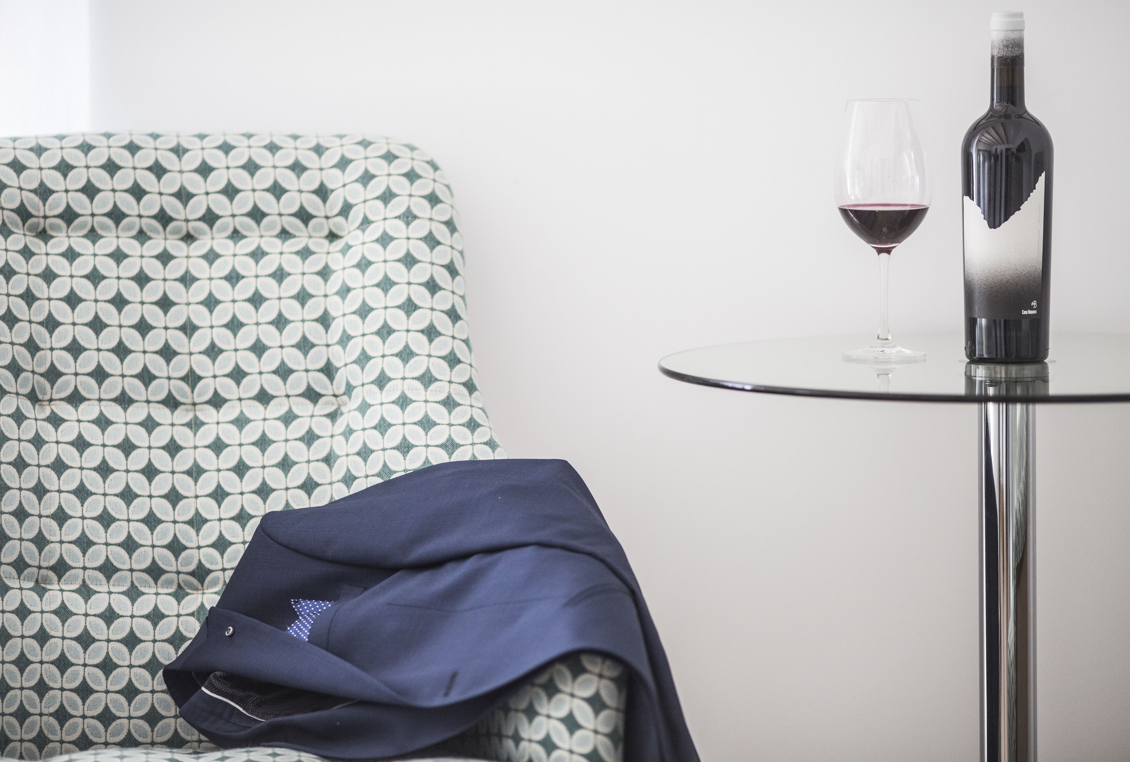 Un sillón con estampado, una mesa con una botella de vino y una copa, y una chaqueta sobre el sillón.