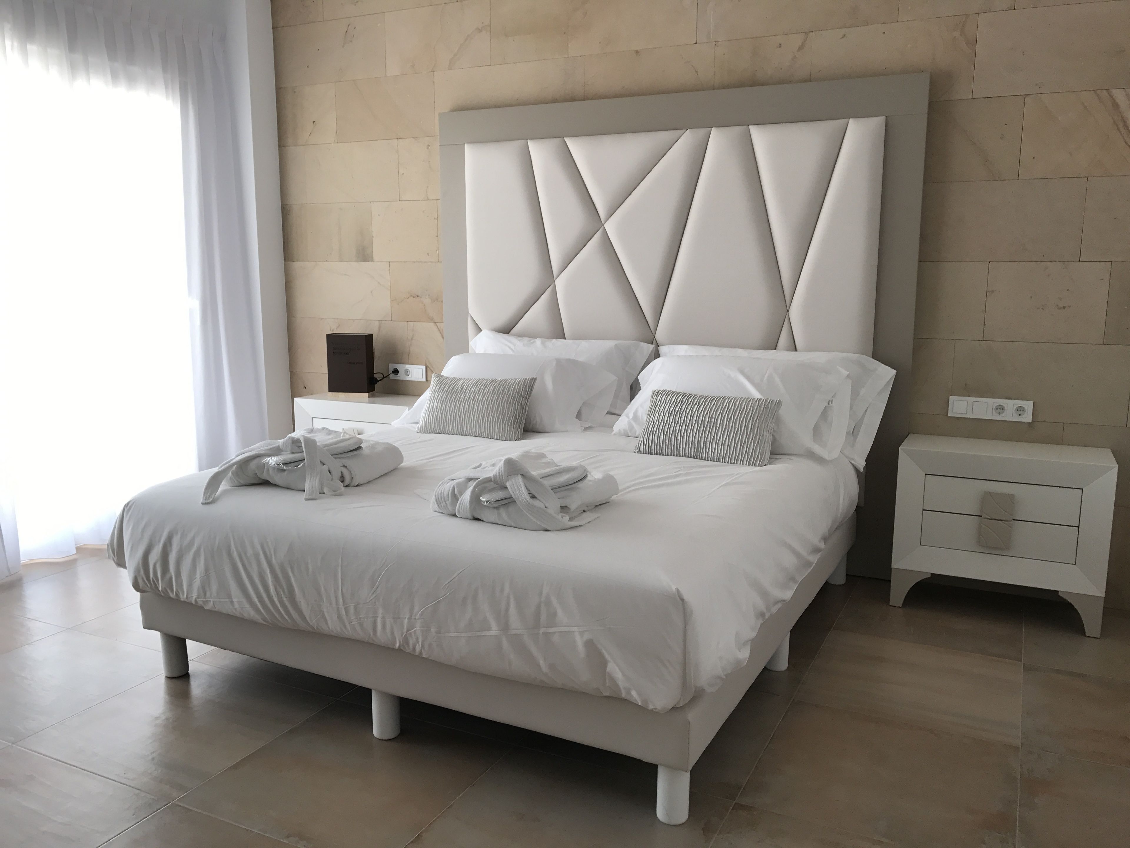 Una habitación de hotel moderna con una cama grande, ropa de cama blanca y un cabecero con diseño de rombos. Hay dos mesitas de noche con lámparas y una mesita auxiliar con una lámpara.