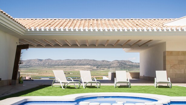 Zona de piscina con vistas a montañas, sillas reclinables blancas y techo de baldosas.