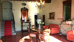 Un comedor con mesa de madera, sillas y una araña. Se observa una escalera con alfombra roja que conduce a un nivel superior.