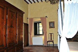 A room with a large wooden wardrobe, a radiator, a chair, and a window with curtains.
