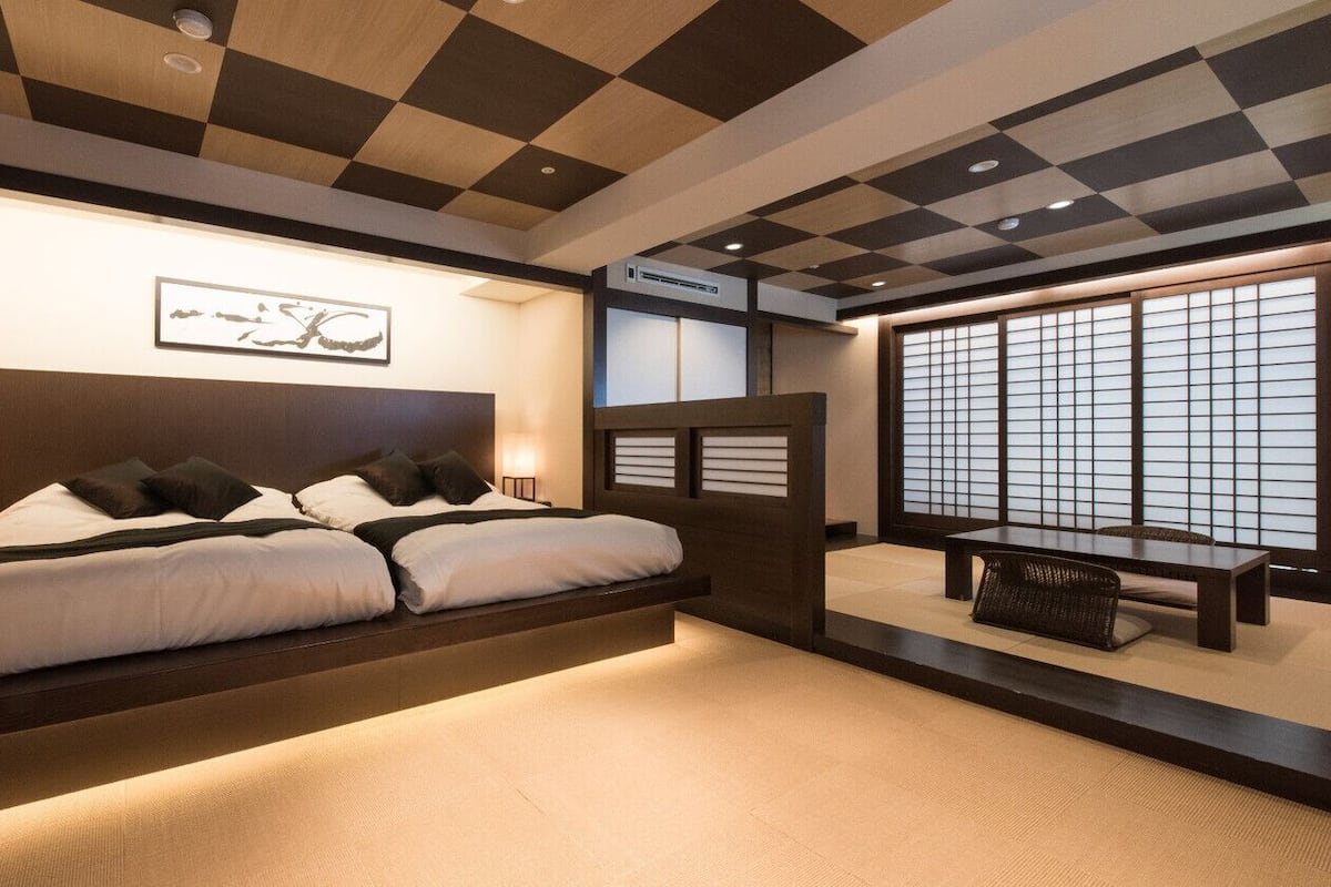 A modern bedroom with a large bed, bedside lamps, a wooden headboard, tatami flooring, and a view of the outside through shoji screens.