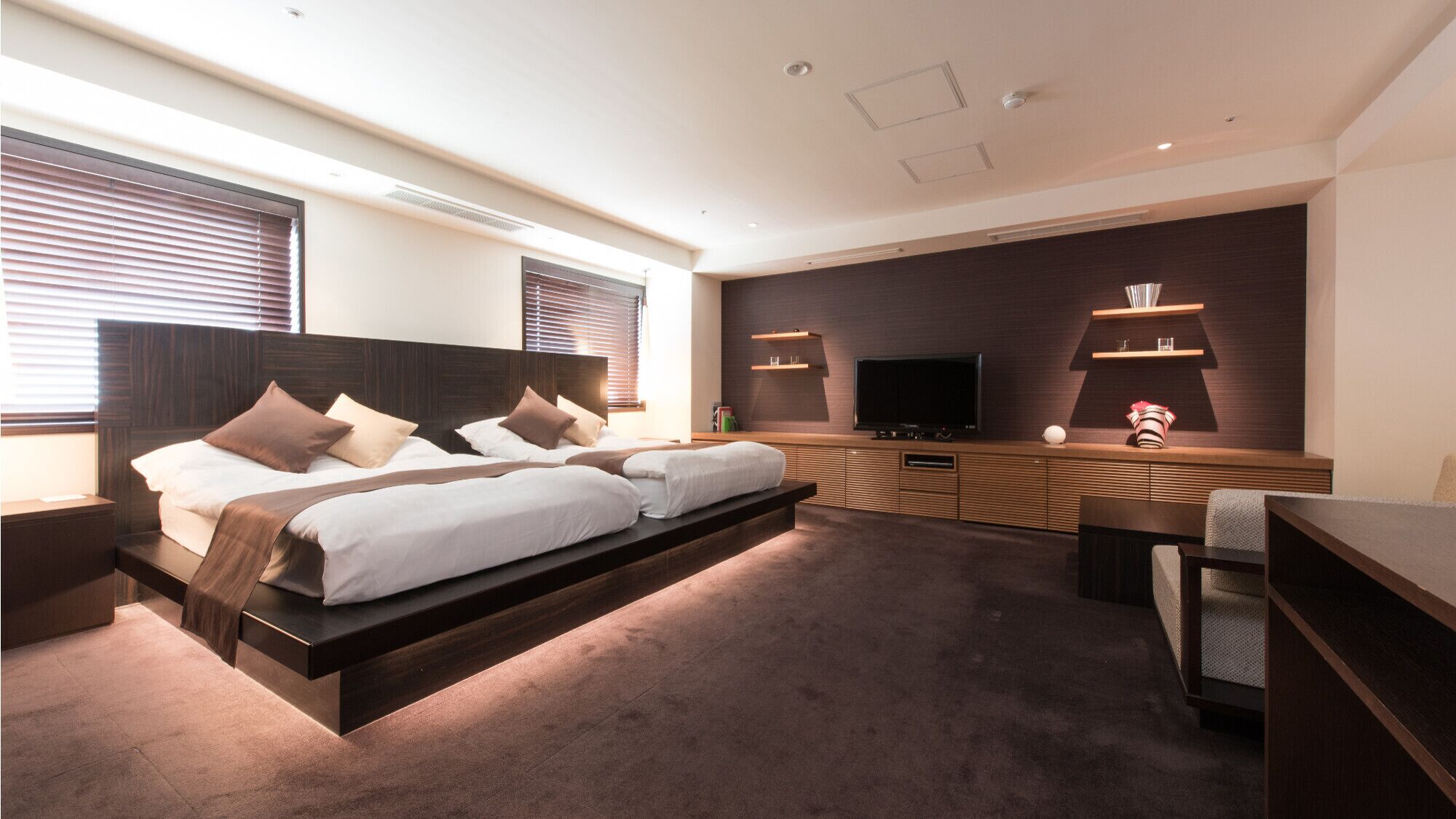 A modern hotel room with a large bed, a flat-screen TV, a wooden desk, and built-in shelving.