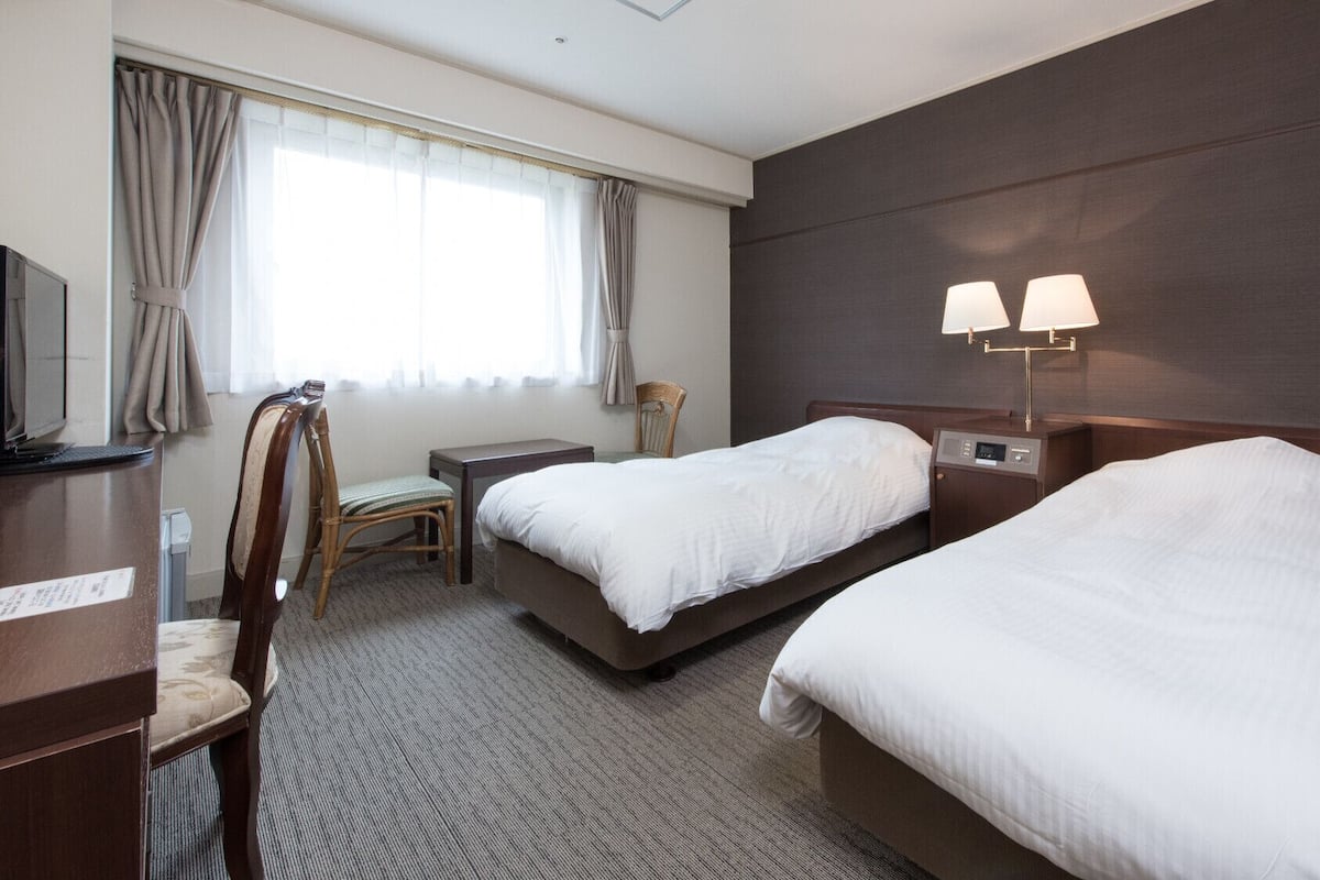 A hotel room with two beds, a desk with a computer, a chair, a lamp, and a window with curtains.