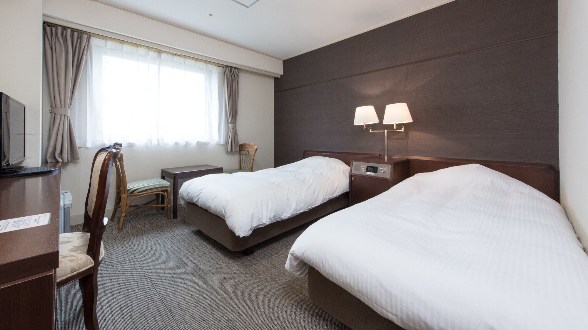 A hotel room with two beds, a desk, a chair, a TV, and a lamp.