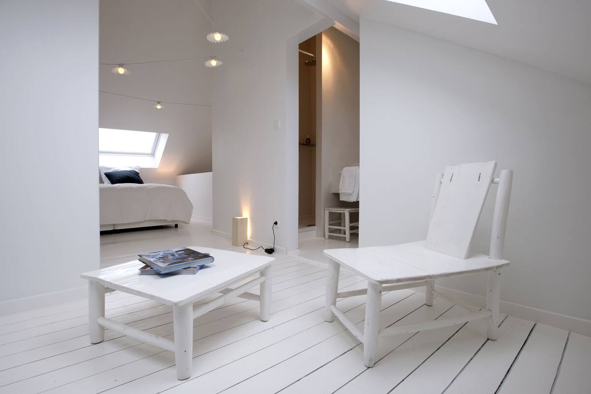 A white room with a bed, a small table with magazines, and a white chair.