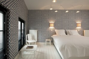 A bedroom with a patterned wallpaper, a bed, a small table, a chair, and two wall-mounted lights.