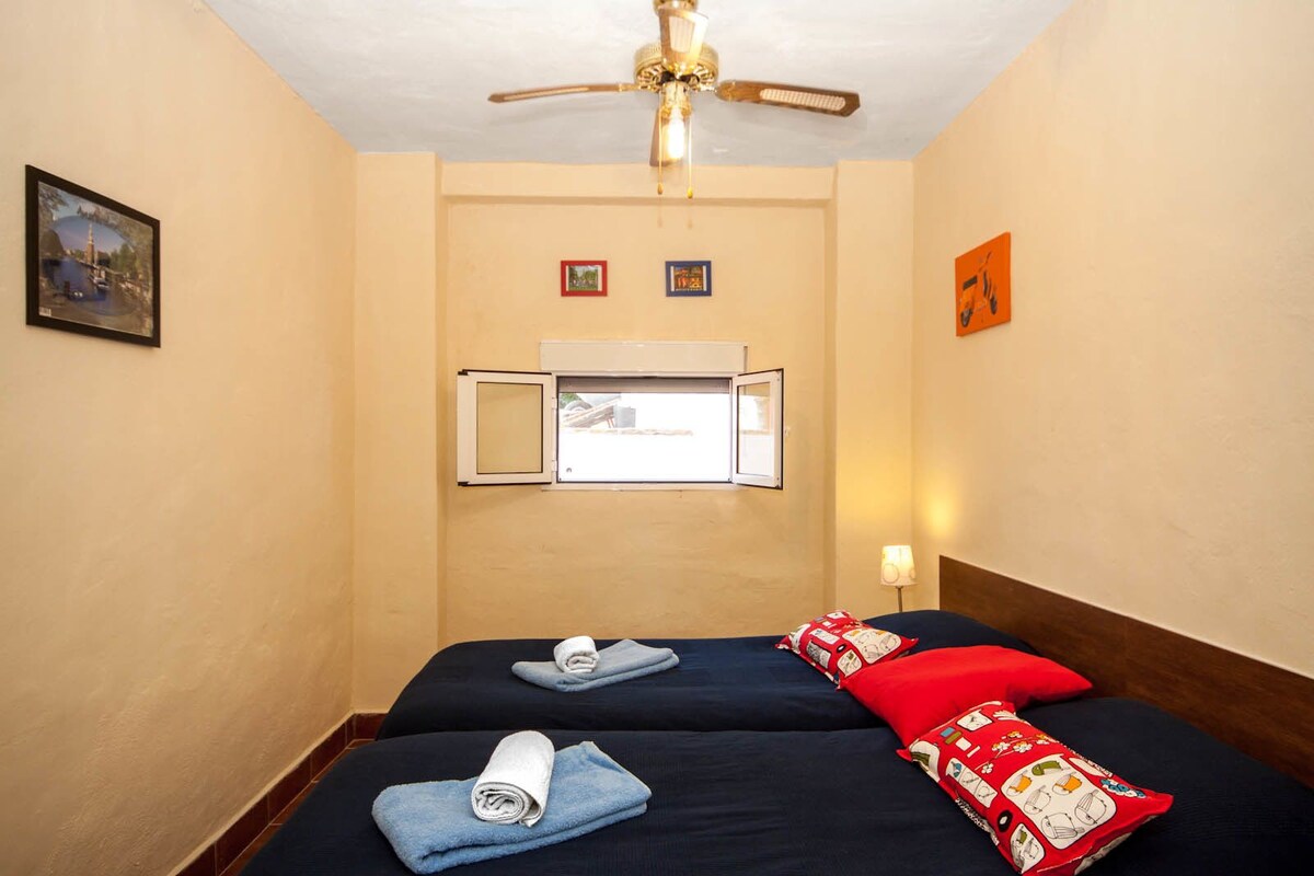 Une chambre d’hôtel avec deux lits, un ventilateur de plafond, une fenêtre avec un appareil de climatisation et un téléviseur fixé au mur.