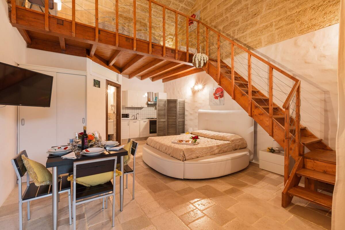 Una stanza con una scala in legno, un letto rotondo, un'area pranzo con tavolo e sedie, e una cucina sullo sfondo.