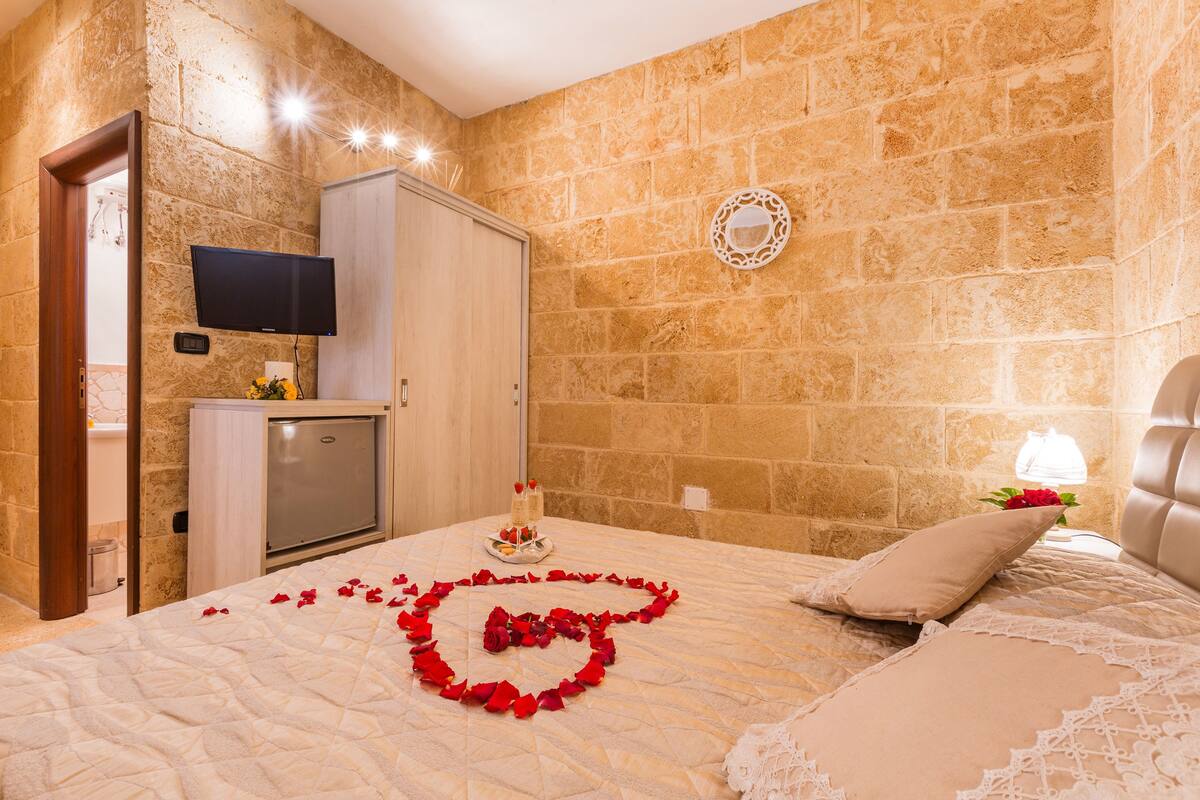 Una camera d'albergo con parete in pietra, un letto con composizione floreale a forma di rosa e un angolo bar, oltre a una televisione.
