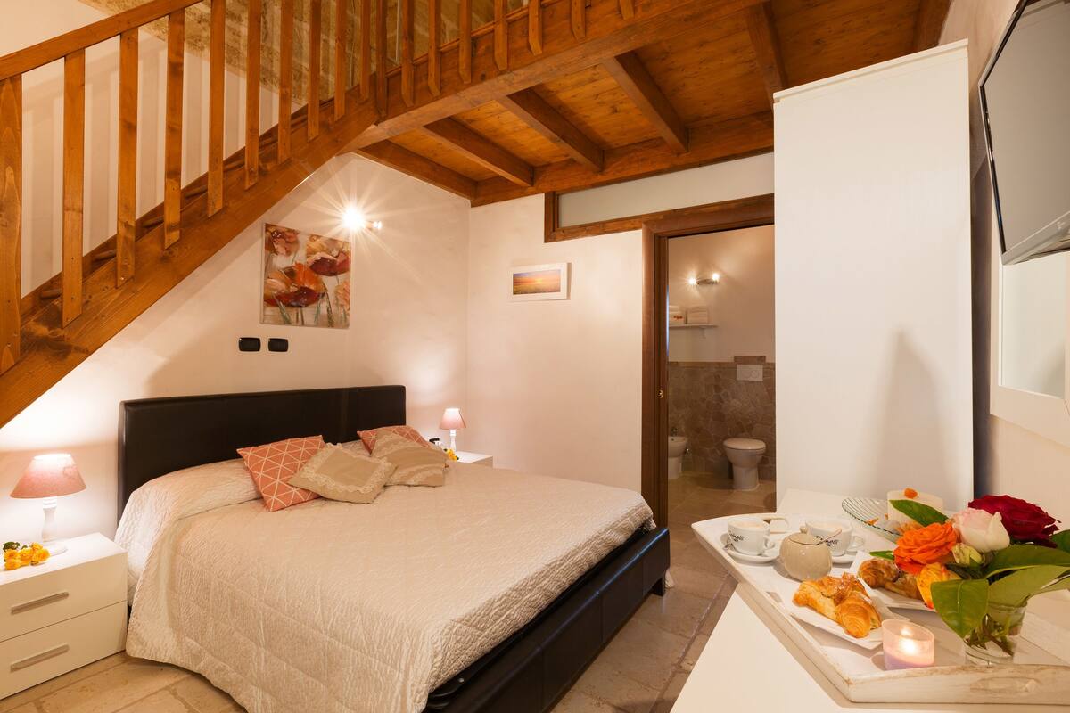 Una camera da letto con una scala in legno, un letto con copripiumino bianco, un comodino con lampada e un vassoio per la colazione con pasticcini e fiori.