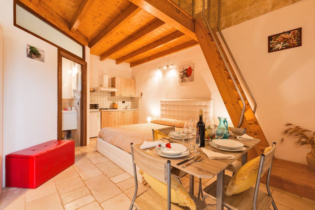Una camera accogliente con soffitto in legno, un letto, un tavolo da pranzo apparecchiato con piatti e bicchieri, e una scatola portaoggetti rossa.