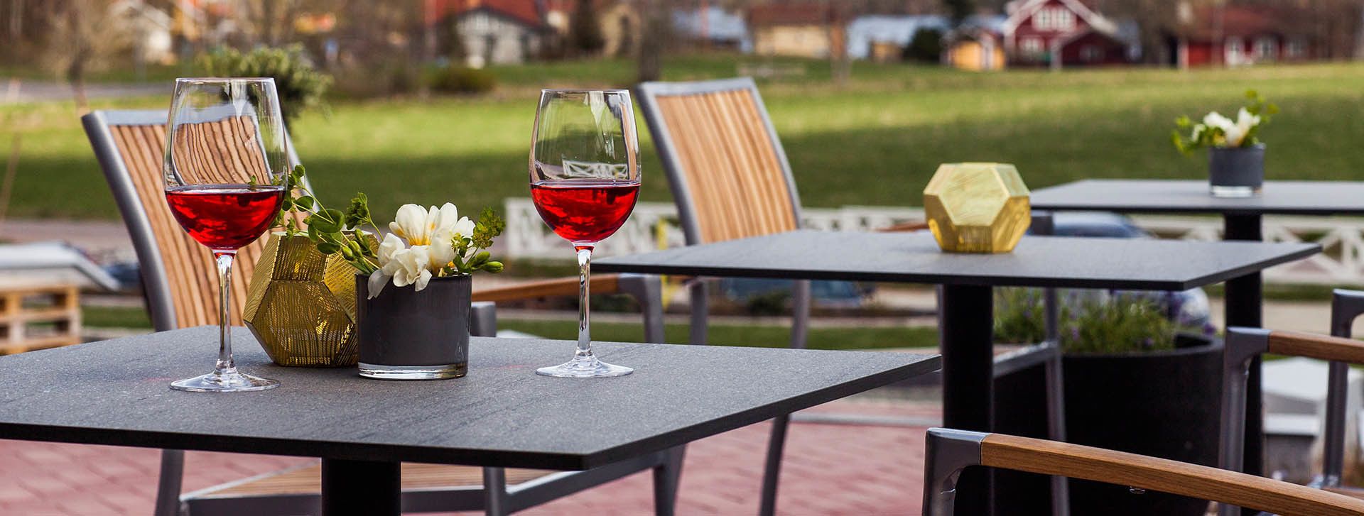 Uma área externa com mesas e cadeiras, dois copos de vinho tinto e um pequeno vaso com flores brancas.