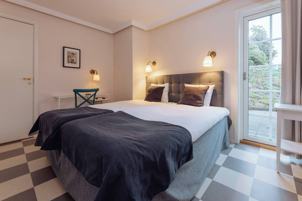 A bedroom with a checkered floor, a bed with dark bedding, two bedside lamps, and a window with a view of greenery.