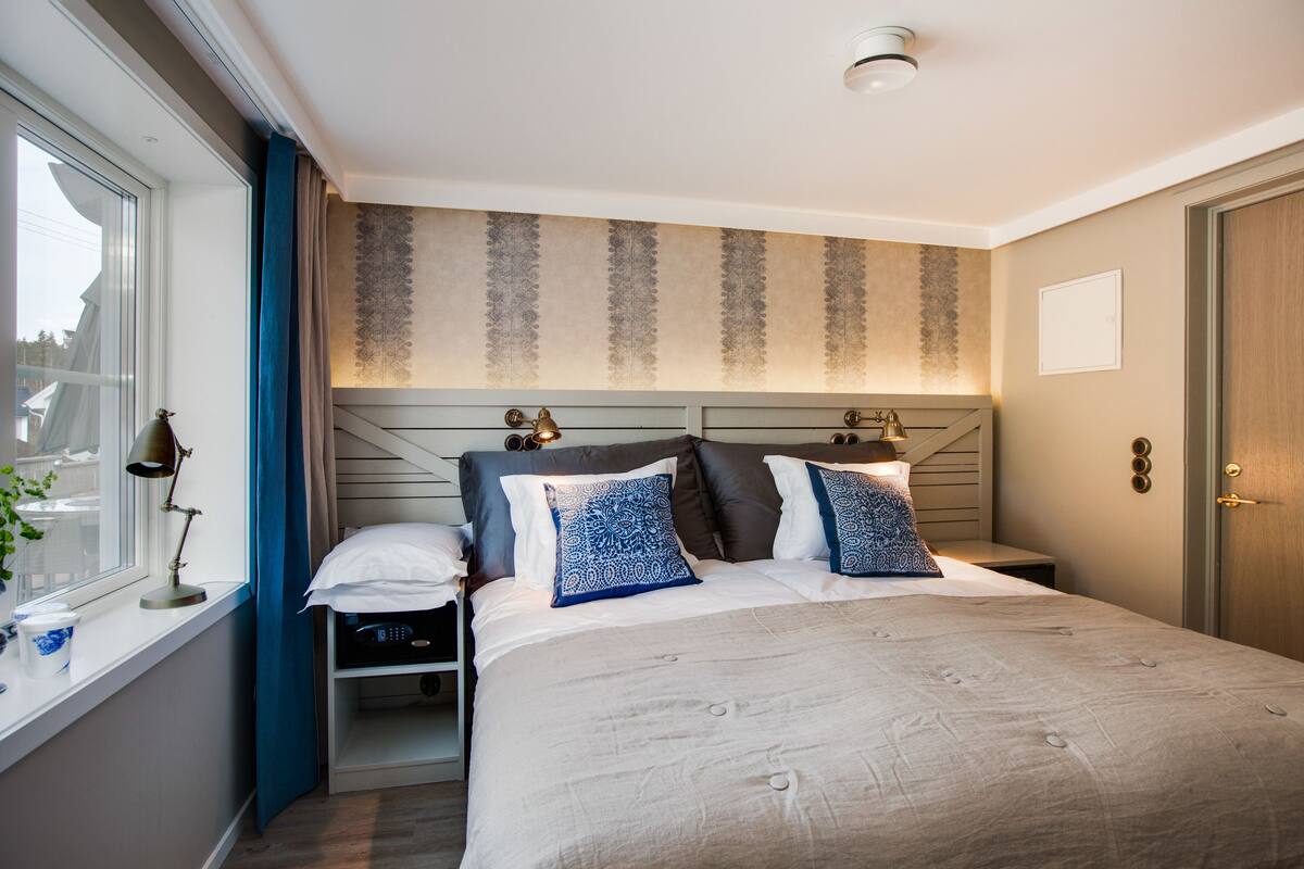 A neatly made bed with a headboard, a bedside table with a lamp, and a window with curtains.