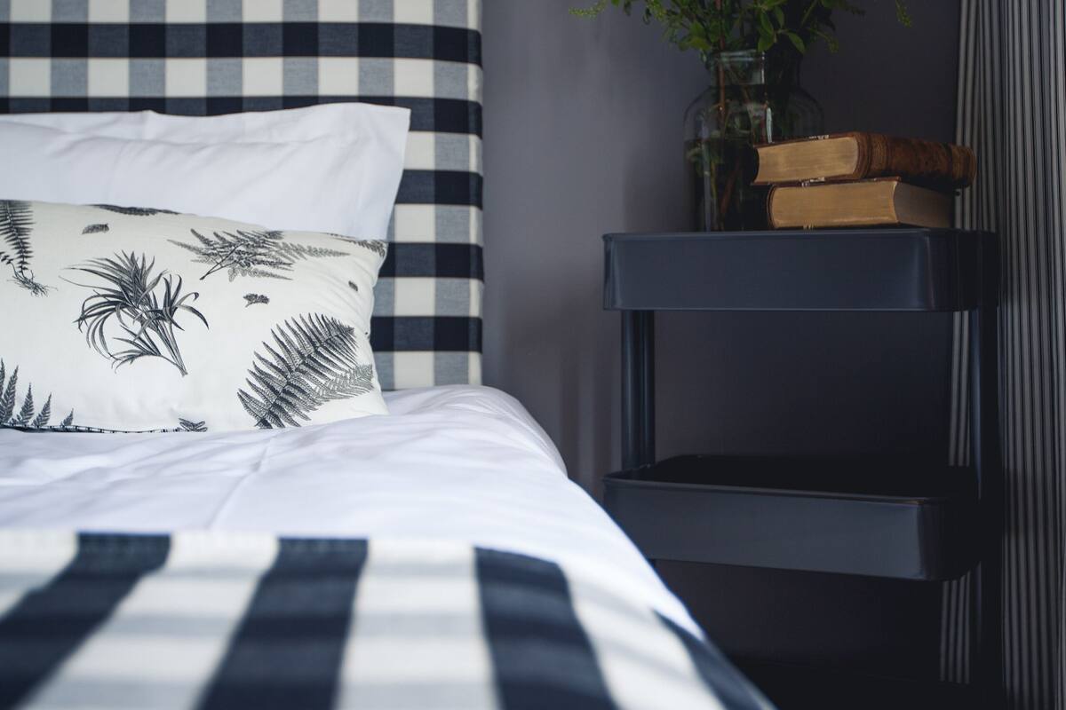 A bed with a checkered headboard, a bedside lamp, a small plant, and a stack of books on a nightstand.