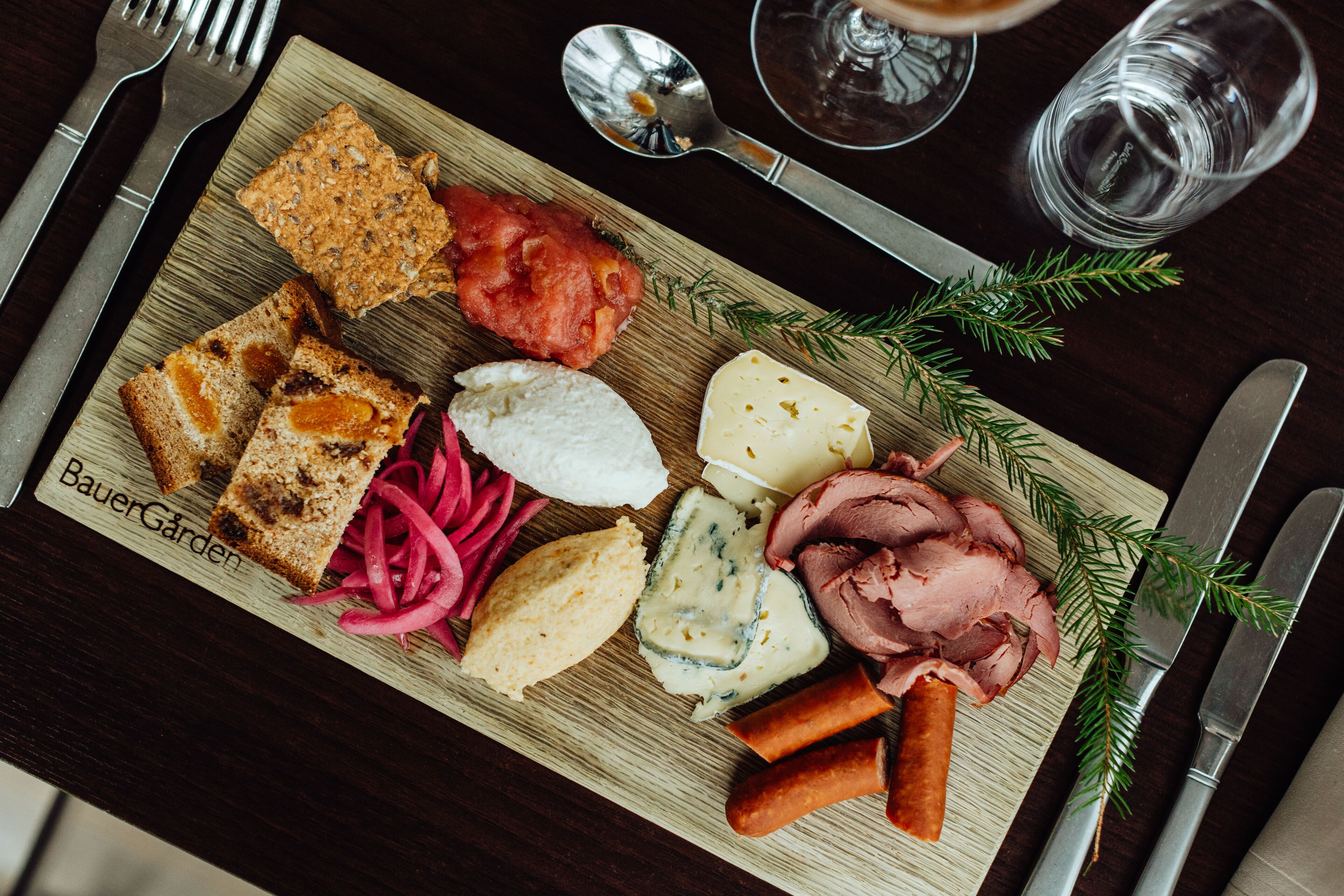 A wooden board with various food items including bread, cheese, and meat, accompanied by cutlery and a glass of water.