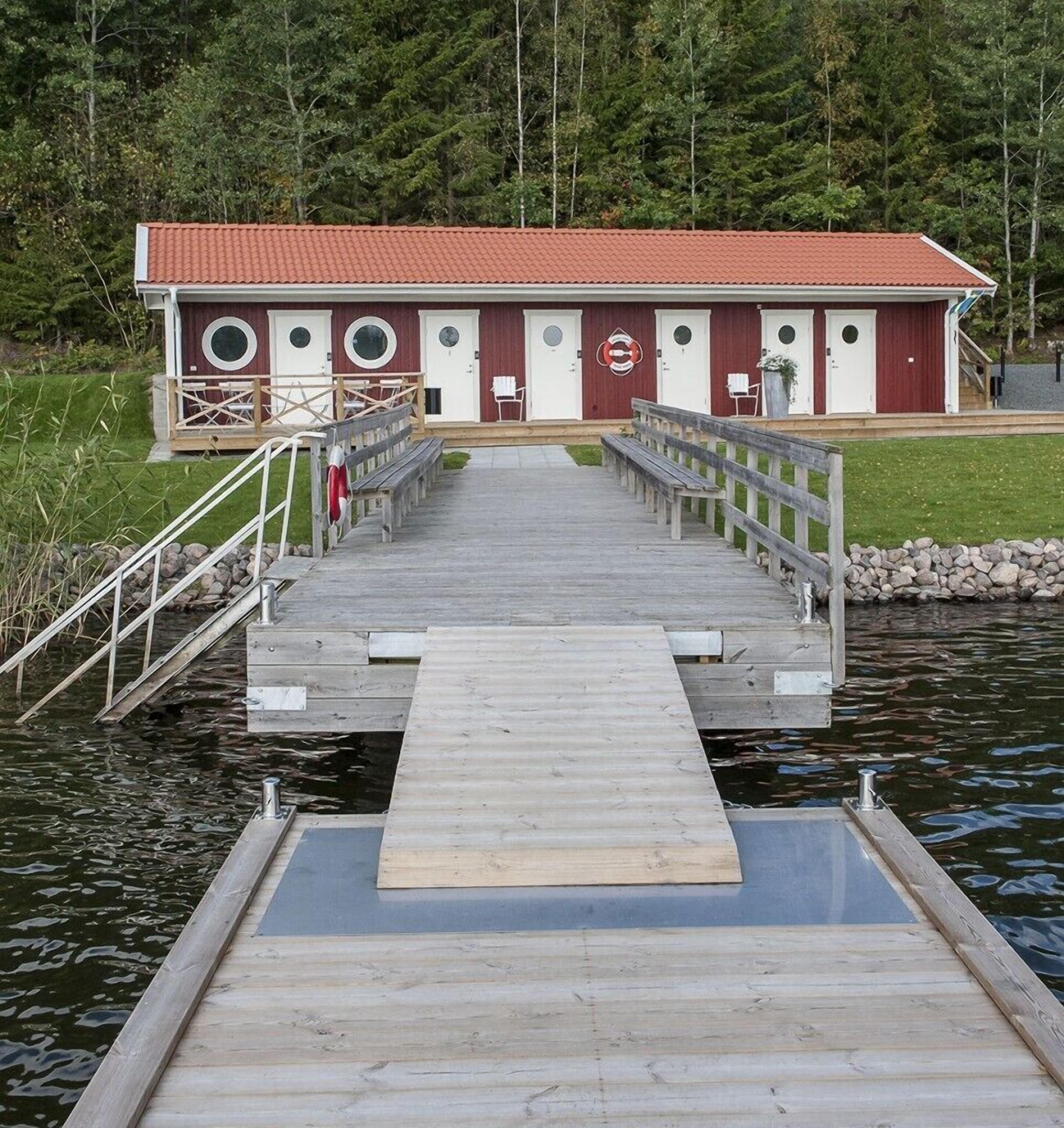 A wooden dock leading to a building with a red roof and white doors, surrounded by water and trees.
