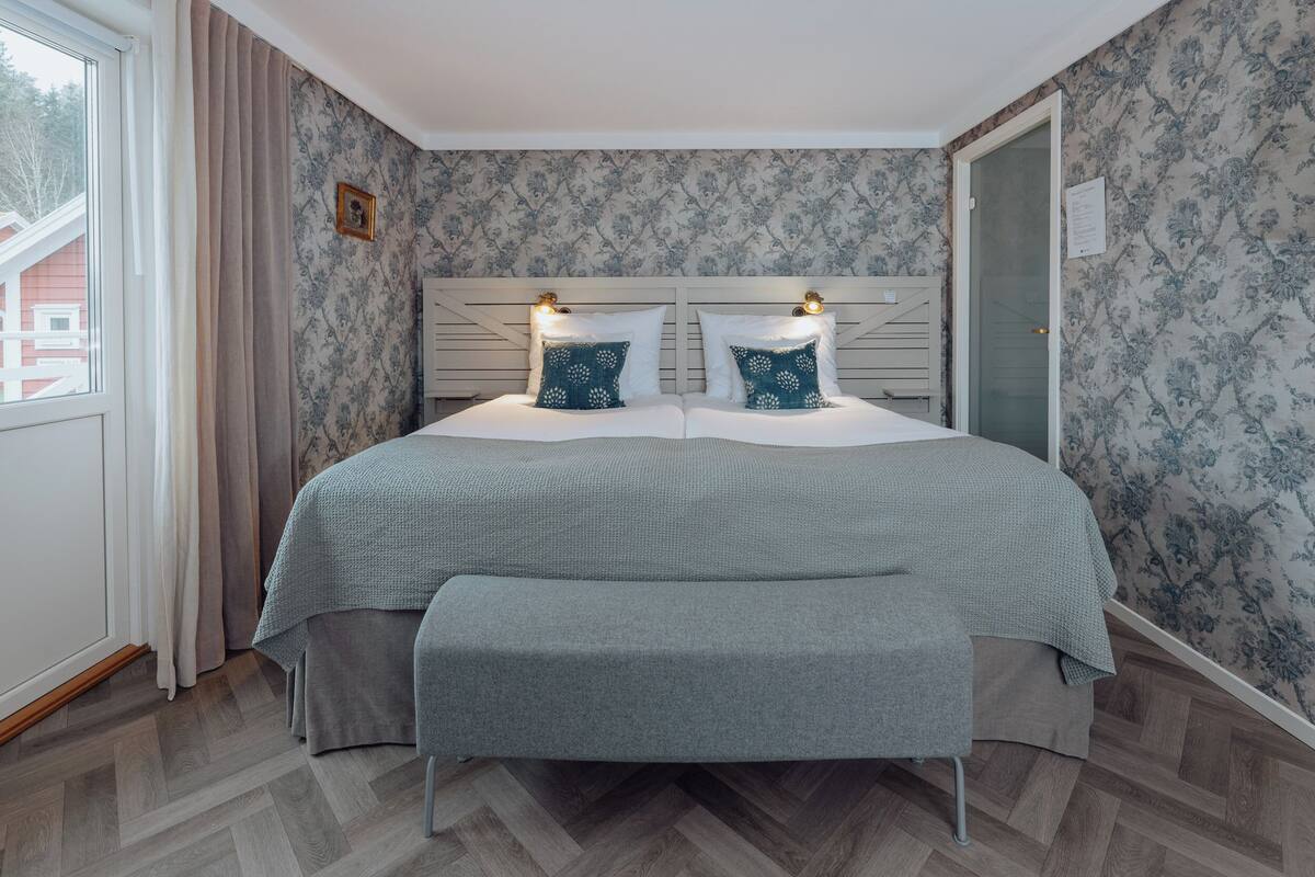 A bedroom with a large bed, two bedside lamps, a bench, and a window with curtains.
