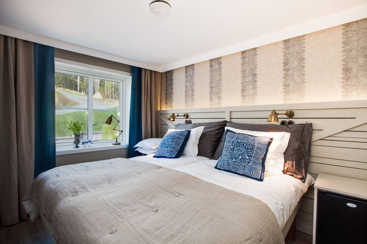 A bedroom with a large bed, a nightstand, a window with curtains, and a view of a green landscape.