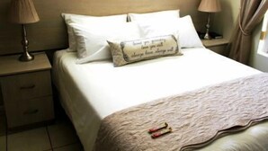 A neatly made bed with a decorative pillow, a bedside table with a lamp, and a window with curtains. - 6 Nendaz Self catering (Langebaan)