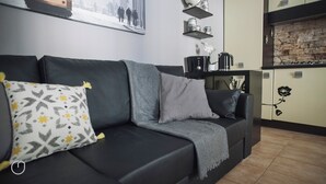 A modern living room with a black sofa, grey cushions, and a patterned pillow. There is a kitchen area with a stone backsplash and a coffee maker on the counter. - Italianway   - San Gottardo (Milan)