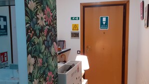 Une pièce dotée d’une porte en bois signalée comme issue de secours, d’un panneau mural indiquant l’emplacement d’un poste de premiers secours, et d’une petite table sur laquelle se trouvent une lampe et des livres.