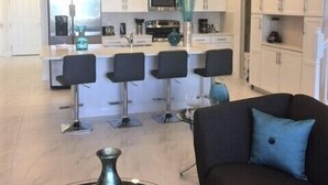 Een moderne keuken met witte kasten, een centraal eiland met barkrukken, en een eethoek met een zwarte bank en decoratieve vazen.