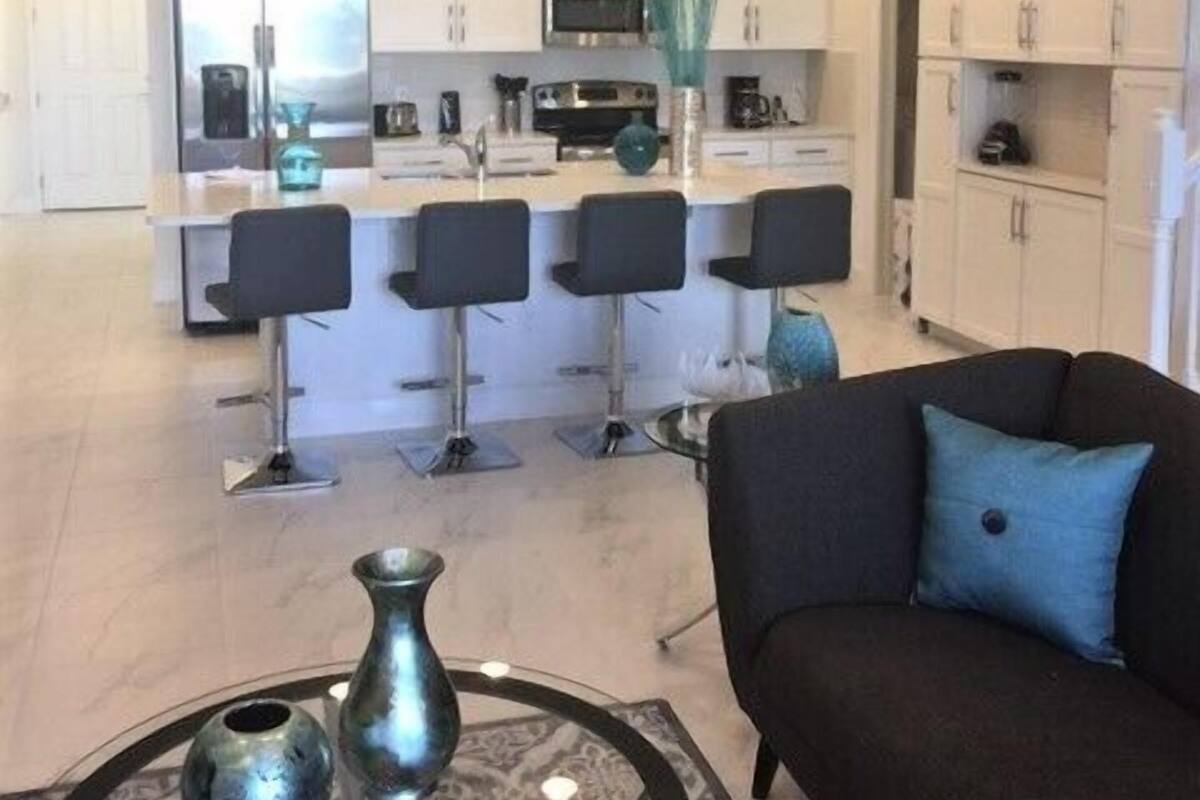 A modern kitchen with white cabinets, a central island with bar stools, and a dining area with a black sofa and decorative vases.