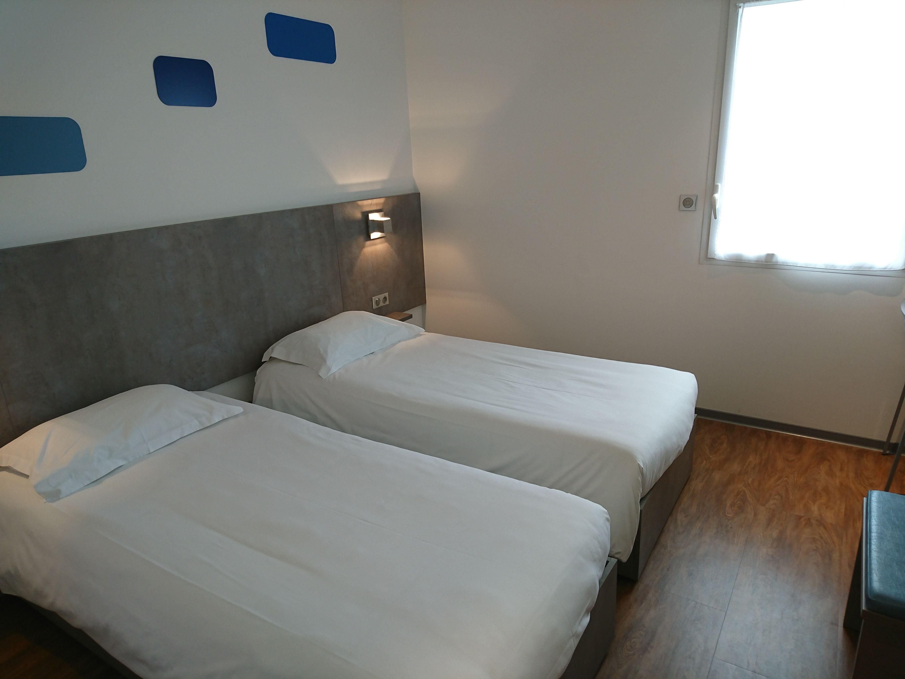 A hotel room with two beds, a wooden floor, and a window with natural light.