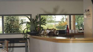 A modern kitchen with a curved counter, a potted plant, and a view of the outdoors through large windows. - H24 HOTEL Le Mans (Le Mans)